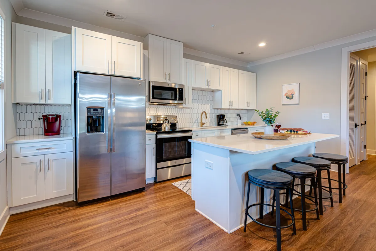 A modern kitchen with a prominent stainless steel refrigerator, a kitchen island with seating, and sleek white cabinetry. The island provides additional space for meal prep or casual dining, and the overall design is clean, bright, and welcoming, ideal for group cooking or sharing meals.