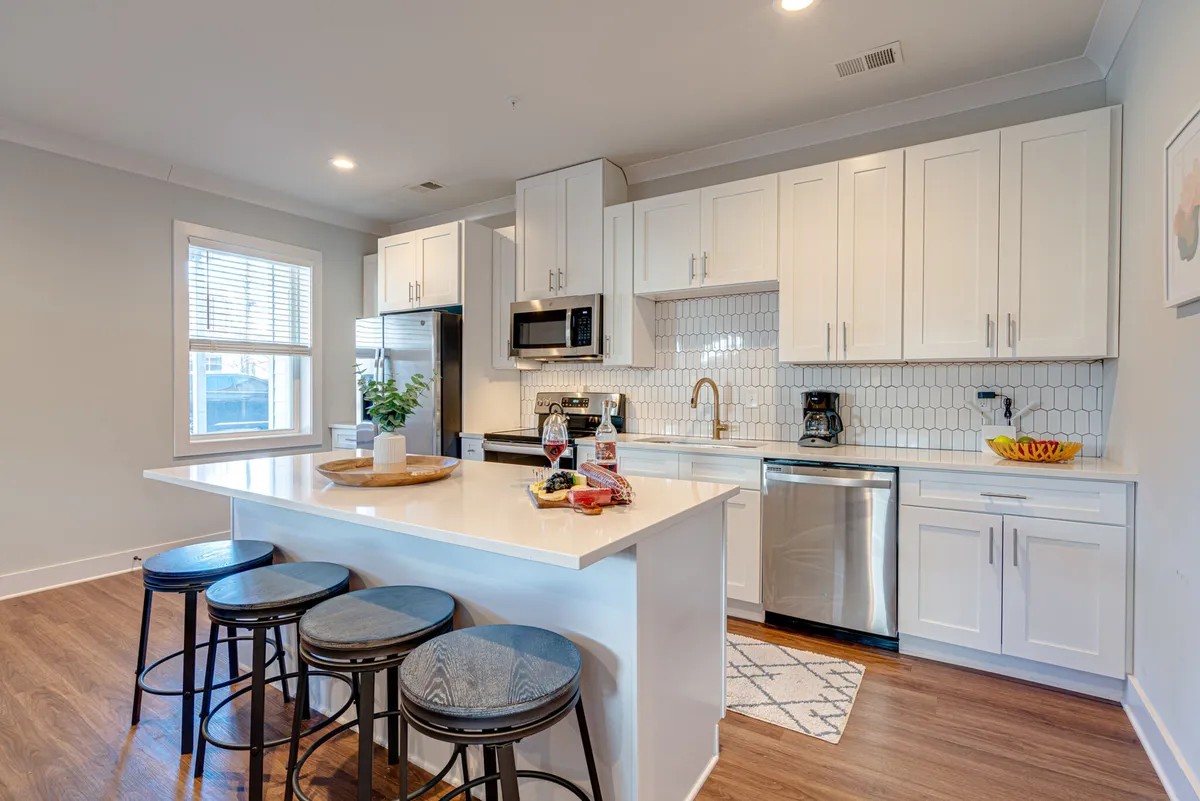 The image showcases a sunlit, modern kitchen with a spacious island, gleaming stainless steel appliances, crisp white cabinetry, a cheerful bowl of fresh fruit, and a decorative rug. The warm and open layout invites social cooking, brunches, and casual gatherings, setting a welcoming tone for the home.