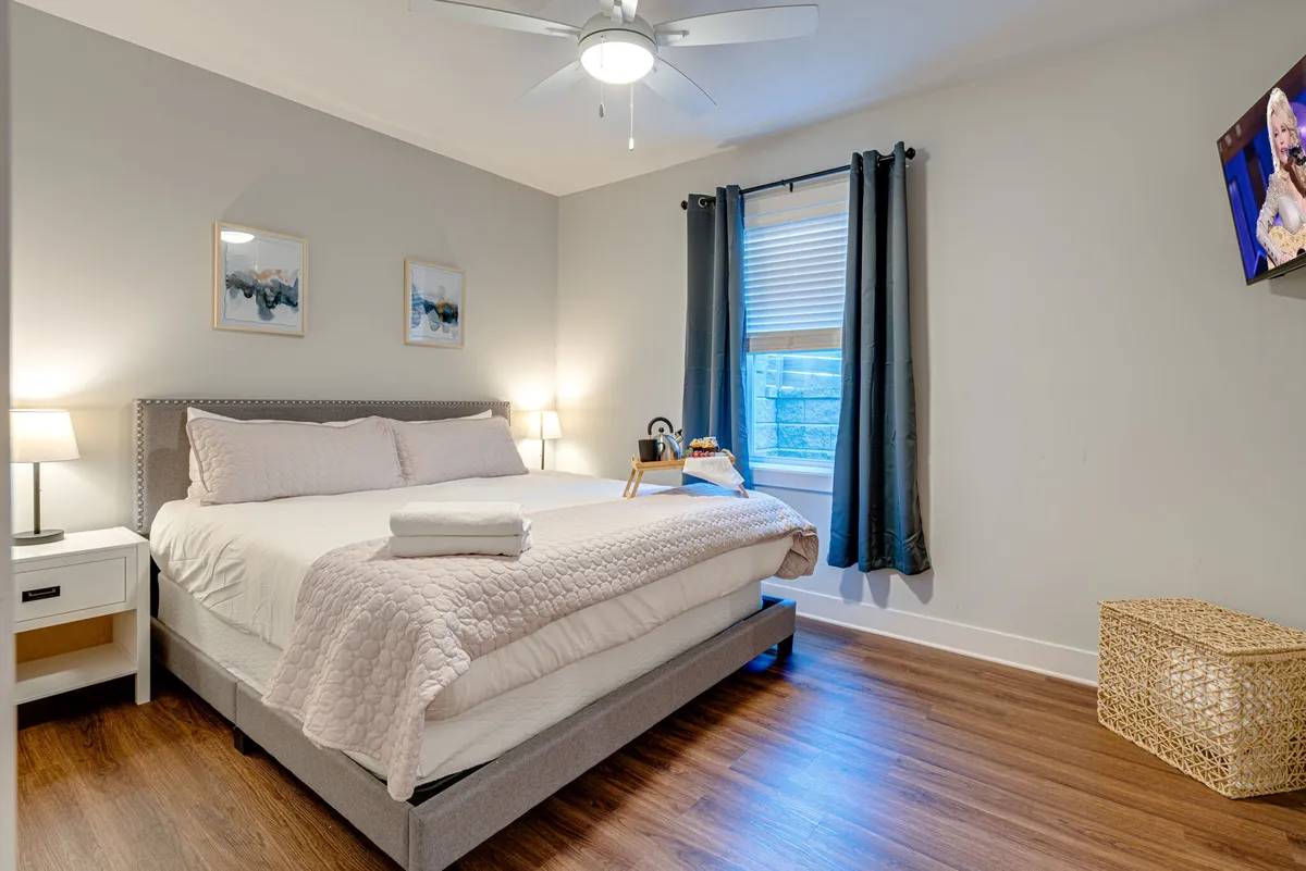 This image features a king-sized bed adorned with decorative pillows, a nightstand with a lamp, and a window with curtains. The space is bright with natural light and has a relaxing, inviting ambiance, ideal for winding down after a busy day. The presence of a TV is implied, perfect for watching shows from bed.