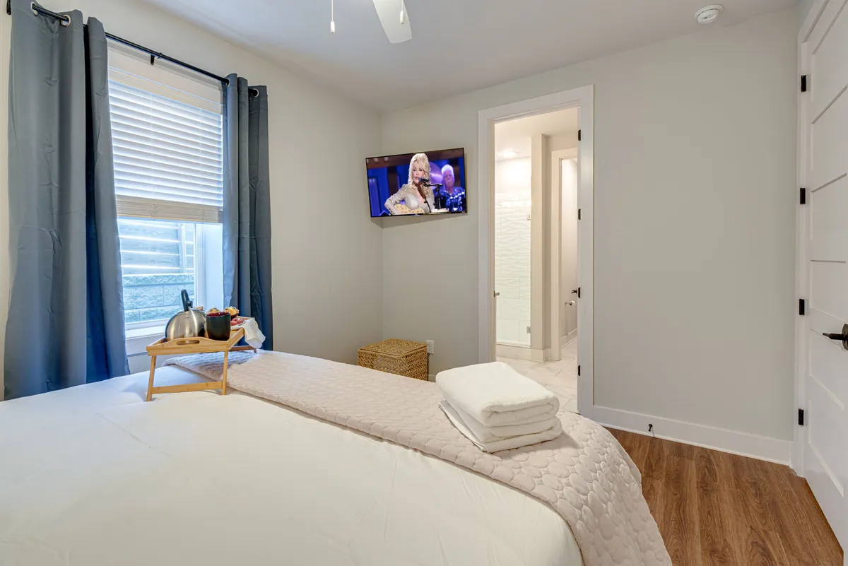 This bedroom scene highlights a bed with neatly arranged linens, a wall-mounted television, and a window with curtains. The setting is cozy and homely, ideal for watching TV or relaxing. The soft décor and thoughtful details evoke the restful retreat described in the section.