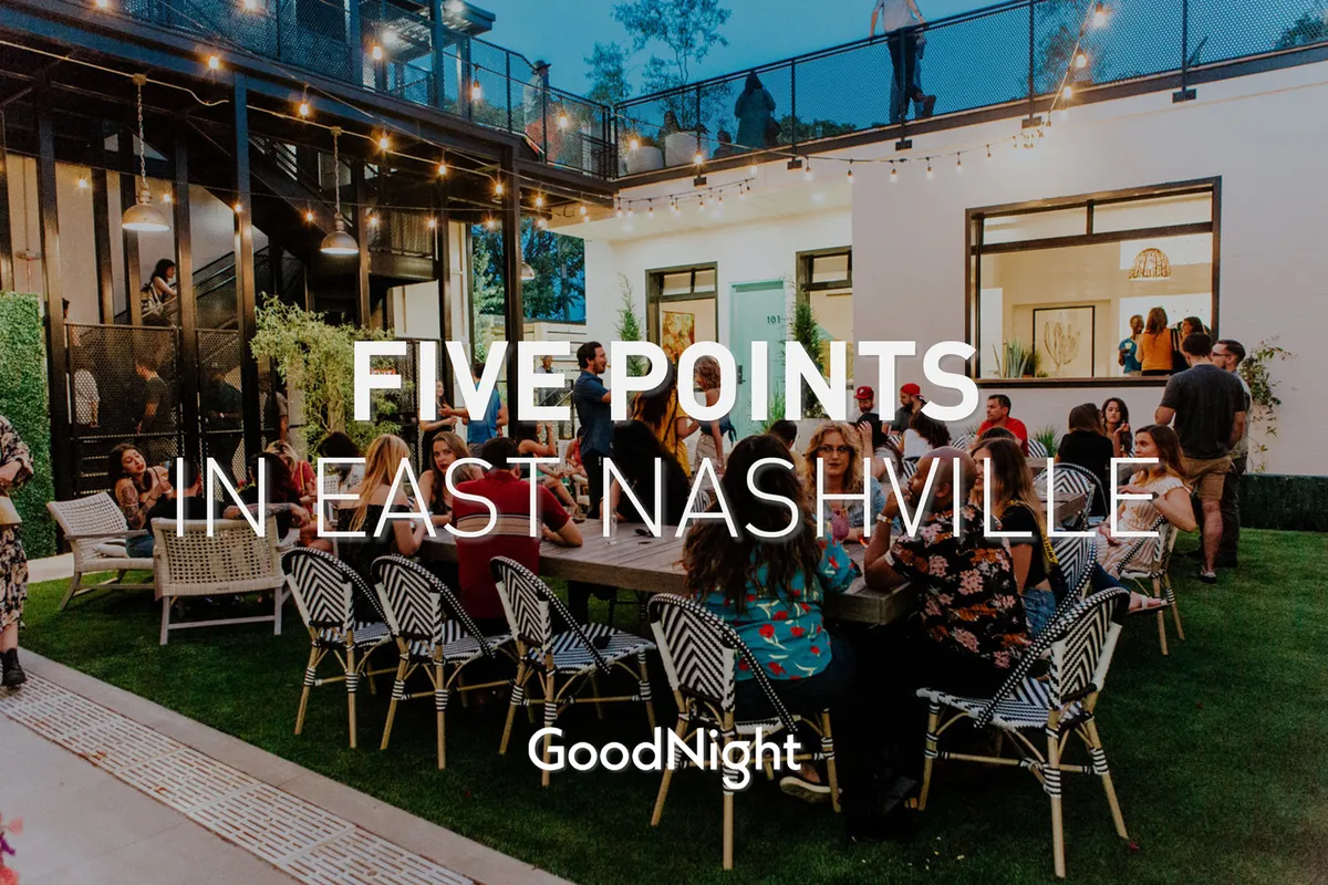 A modern outdoor space featuring a large communal table lit by string lights, with people dining and socializing under an open sky. The contemporary architecture and festive setting suggest a lively, community-focused atmosphere.