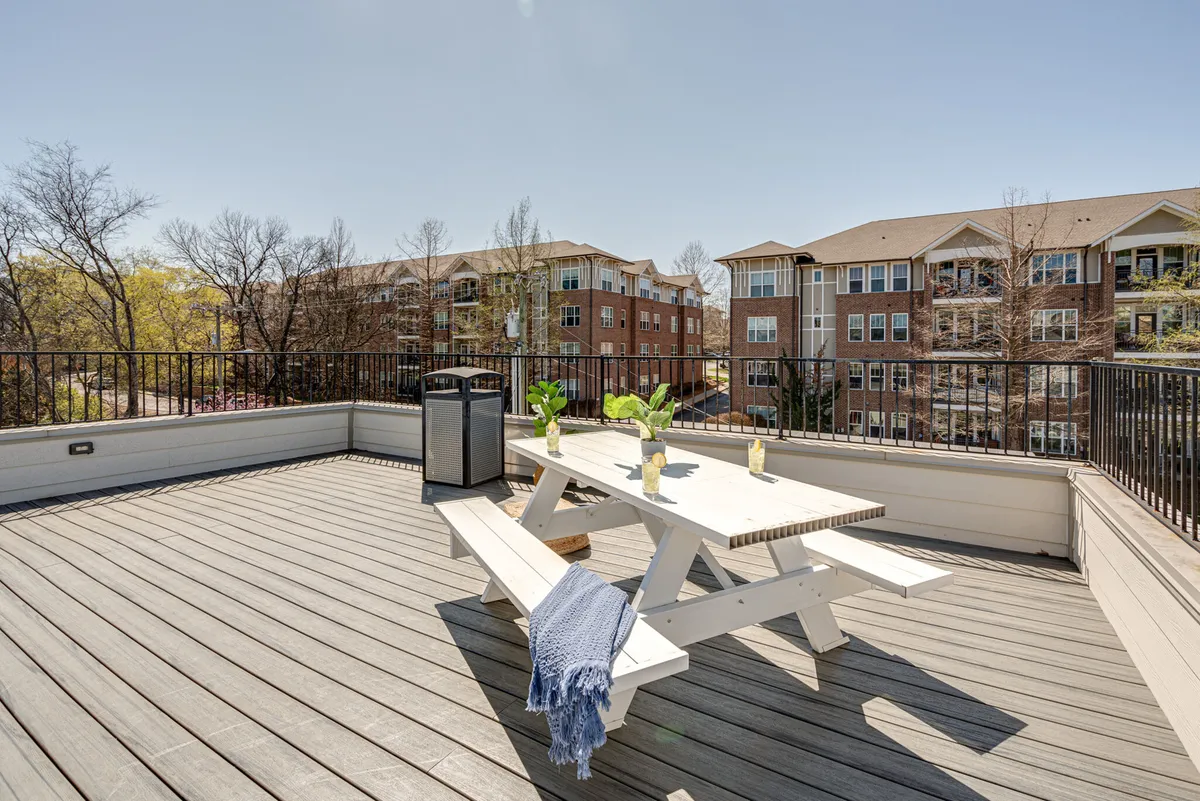 An exterior communal deck scene with a dining table surrounded by lush green plants. The relaxed atmosphere hints at sunset dinners and morning coffees, perfect for small gatherings and socializing in an outdoor setting.