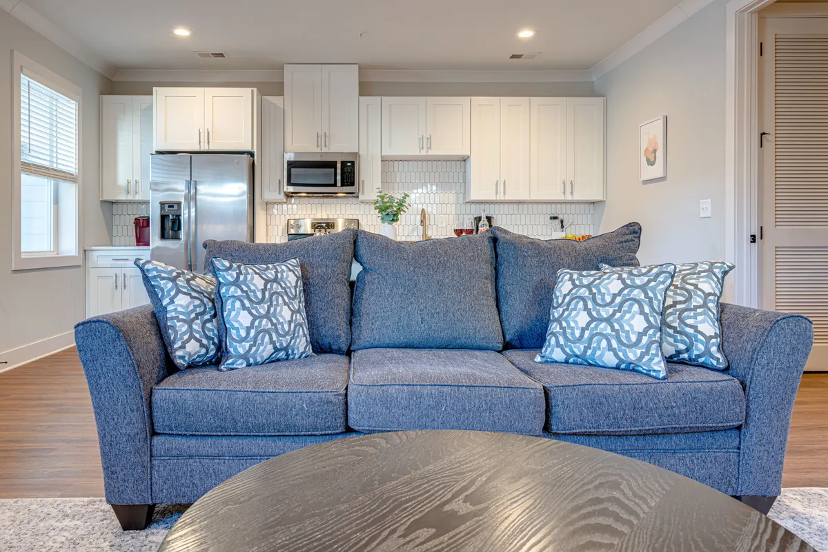 A comfortable, inviting living area featuring a blue couch with decorative pillows and a coffee table, seamlessly connected to a modern kitchen in the background. The open layout allows for easy movement between the living and kitchen spaces, with ample light and stylish decor that balances comfort and sophistication.